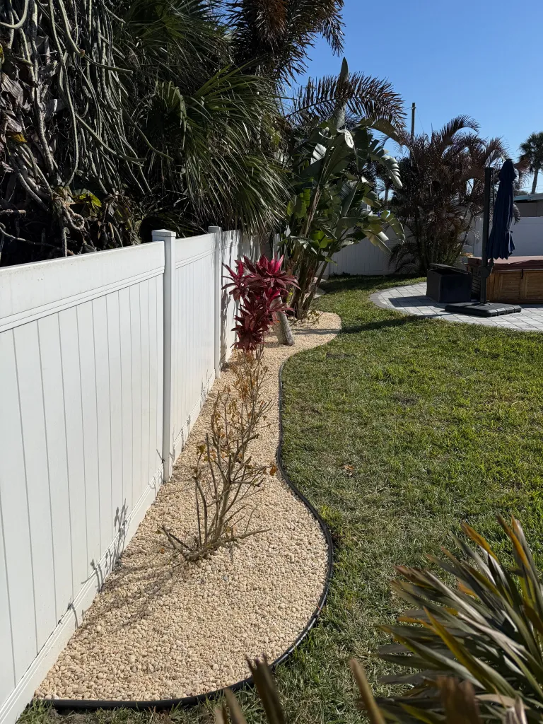 Curved decorative rock bed with tropical plants along a white fence
